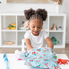 Load image into Gallery viewer, A smiling child with curly hair sits at a white table, reaching into a Bumkins Wet Bag: Disney Princess Ariel while surrounded by toys.
