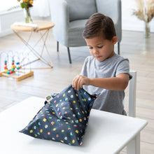 Load image into Gallery viewer, A boy in a gray shirt sits at a white table, opening a Bumkins Wet Bag: Super Mario™ Lineup in a modern room with toys.
