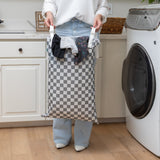 In a laundry room, a person holds a Bumkins Extra Large Wet Bag: Charcoal Check filled with clothes near a washing machine.