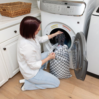 A woman kneels by a front-loading washer, placing clothes from a Bumkins Extra Large Wet Bag: Charcoal Check inside.