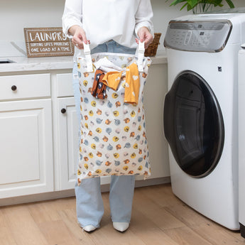 A person holding a Bumkins Extra Large Wet Bag: Winnie + Friends with clothes in a modern laundry room, between a washing machine and cabinet.