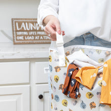 Load image into Gallery viewer, Close-up of a person holding a Bumkins Extra Large Wet Bag: Winnie + Friends filled with clothes in a bright laundry room.
