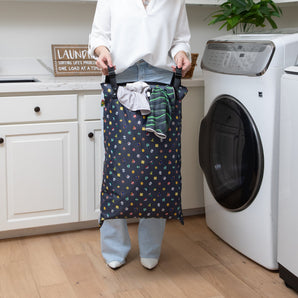 In the laundry room, a person holds a Bumkins Extra Large Wet Bag: Super Mario™ Lineup with snap handles, filled with clothes.