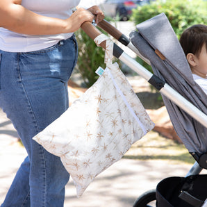 A person pushes a stroller outdoors with a child in it, featuring a Bumkins Wet/Dry Bag: Palm Daze hanging from the handle.