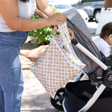 Load image into Gallery viewer, A person places clothes into a Bumkins Wet/Dry Bag: Sand Check attached to a stroller beside a sitting child outdoors on a sunny day.
