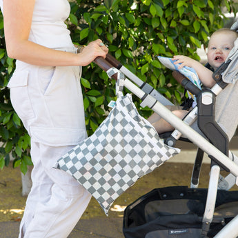A person pushes a stroller holding a baby, with a Bumkins Wet/Dry Bag: Charcoal Check attached, outdoors.