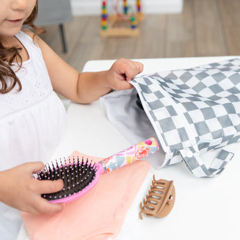A child places a hairbrush and clip into a Bumkins Wet/Dry Bag: Charcoal Check on a white table.