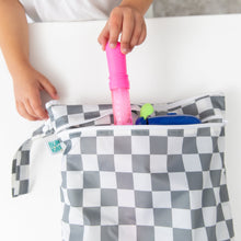 Load image into Gallery viewer, A child places a pink item into a Bumkins Wet/Dry Bag: Charcoal Check, featuring a zipper and waterproof fabric, on a white table.
