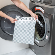 Load image into Gallery viewer, A person holds Bumkins Wet/Dry Bag: Charcoal Check in front of an open front-loading washing machine.
