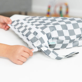 Hands zip a Bumkins Wet/Dry Bag: Charcoal Check on a white table.
