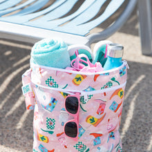 Load image into Gallery viewer, Bumkins Wet/Dry Bag: Boots holds a towel, water bottle, swimwear, and sunglasses in front of an empty lounge chair.
