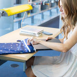 In a library, a woman at a desk pulls a laptop from a Bumkins Wet/Dry Bag: Evil Eye, with books and a lamp next to her.