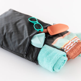 Roamio by Bumkins Wet/Dry Bag: Rich Black holds a light blue towel, swim trunks, toy shovels, & teal sunglasses on a white background.