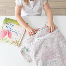 Load image into Gallery viewer, A child fills a Bumkins Wet/Dry Bag: Wander with treasures, while a colorful book sits nearby on a white table.
