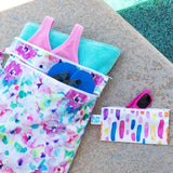 Bumkins Wet/Dry Bag: Watercolor lays next to a pool, holding flip flops, a swimsuit, and a towel; a small bag with sunglasses sits nearby.