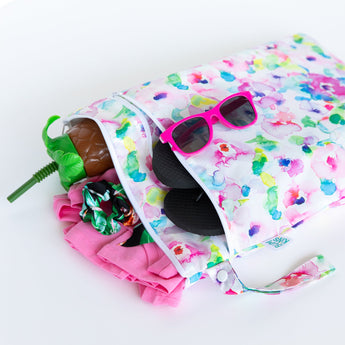 Bumkins Wet/Dry Bag: Watercolor holds pink sunglasses, a toy pineapple, and clothes, all on a white background.