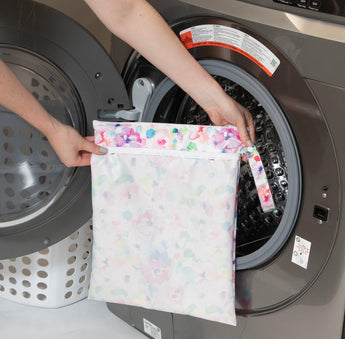 A person holds Bumkins Wet/Dry Bag: Watercolor in front of an open front-loading washing machine.