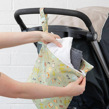 Load image into Gallery viewer, A person places clothing into a Bumkins Wet/Dry Bag: Camp Gear attached to a stroller handle with a white brick wall in the background.
