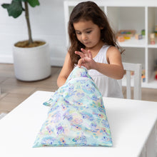Load image into Gallery viewer, A girl in a white dress opens a colorful Bumkins Wet/Dry Bag: Aloha Stitch at a white table in a bright room with plants and shelves.
