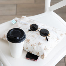 Load image into Gallery viewer, Sunglasses and a smartphone rest on a Bumkins Wet/Dry Clutch: Palm Daze next to a white coffee cup with a black lid on a white chair.
