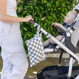An adult with a baby in a stroller, featuring a Bumkins Wet/Dry Clutch: Charcoal Check attached to the handle, amidst lush greenery.
