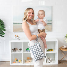 Load image into Gallery viewer, A smiling woman holds a baby and a Bumkins Wet/Dry Clutch: Charcoal Check in a bright room with toys on a shelf.

