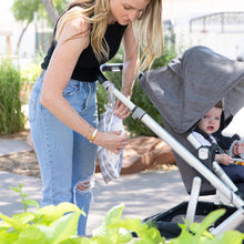 Load image into Gallery viewer, A woman in jeans and a black top places items into a Bumkins Wet/Dry Clutch: Lunar Phase attached to a stroller with her child.
