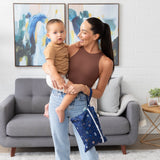 A smiling woman holds a toddler in a living room with paintings and a gray couch, carrying a Bumkins Wet/Dry Clutch: Evil Eye on her arm.