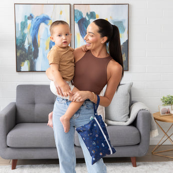 A smiling woman holds a toddler in a living room with paintings and a gray couch, carrying a Bumkins Wet/Dry Clutch: Evil Eye on her arm.