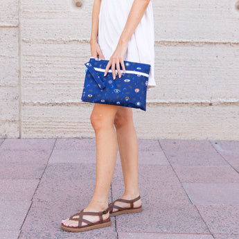 A person in a white dress & sandals holds a Bumkins Wet/Dry Clutch: Evil Eye on tiled pavement against a beige textured wall.