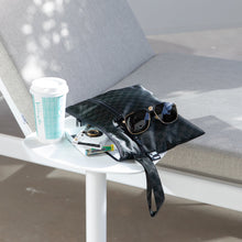 Load image into Gallery viewer, Roamio by Bumkins Wet/Dry Clutch: Rich Black sits on a table outdoors next to a lounge chair with sunglasses, books, and a coffee cup.
