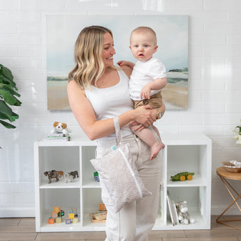 A woman smiles holding her baby in a room with white shelves and toys, carrying a Bumkins Wet/Dry Clutch: Wander.