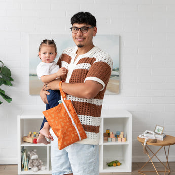 In a living room, a man holds a child and a  Bumkins Wet/Dry Clutch: Grounded; white shelves and wall art are in the background.