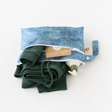 Load image into Gallery viewer, Roamio by Bumkins Wet/Dry Clutch: Crushed Denim lays open on a white background, holding clothes, a lotion bottle, and brown packet.
