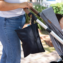 Load image into Gallery viewer, A person pushes a stroller with a baby inside and a Bumkins Wet/Dry Clutch: Black attached to the handle outdoors on a sunny day.
