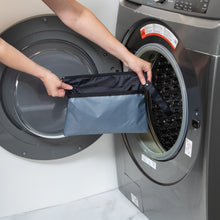 Load image into Gallery viewer, A person holds a Bumkins Wet/Dry Clutch: Black in front of a front-loading washing machine in a laundry room.
