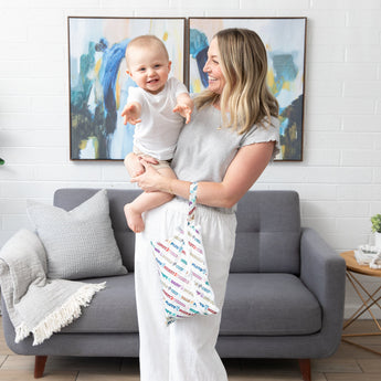 A woman holds a smiling baby and a Bumkins Wet/Dry Clutch: Mickey & Friends Varsity in a cozy living room.
