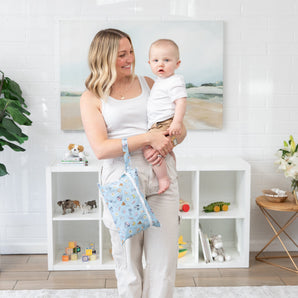 A woman holds a baby in a bright room, surrounded by toys and plants, with a Bumkins Wet/Dry Clutch: Mickey + Minnie Sunny Side Up on her wrist.
