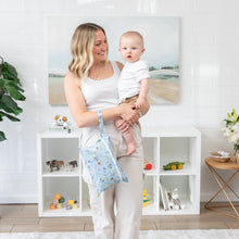Load image into Gallery viewer, A woman holds a baby in a bright room, surrounded by toys and plants, with a Bumkins Wet/Dry Clutch: Mickey + Minnie Sunny Side Up on her wrist.
