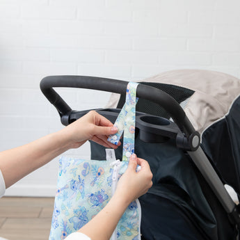 A person attaches a Bumkins Wet/Dry Clutch: Aloha Stitch to a stroller handle against a white brick wall.
