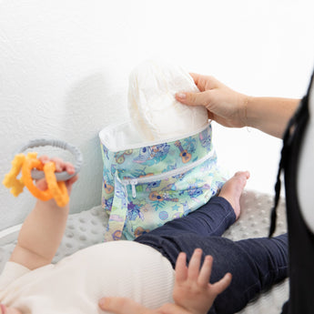 A person pulls a diaper out of a Bumkins Wet/Dry Clutch: Aloha Stitch on a changing table next to a baby holding a teething toy.