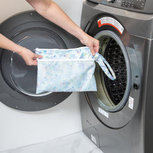 Load image into Gallery viewer, A person holds a Bumkins Wet/Dry Clutch: Aloha Stitch in front of an open front-loading washing machine.
