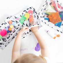 Load image into Gallery viewer, A child reaches into Bumkins Clear Travel Bag 3-Pack: Hearts filled with craft supplies on a white table.
