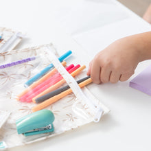 Load image into Gallery viewer, Close-up of a child placing colorful pens in a Bumkins Clear Travel Bag 3 Pack: Palm Daze, with a stapler and sticky notes nearby on a table.
