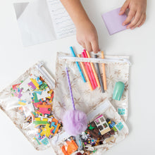 Load image into Gallery viewer, A child's hand reaches for colored pens &amp; toys in Bumkins Clear Travel Bag 3 Pack: Palm Daze on a white table.
