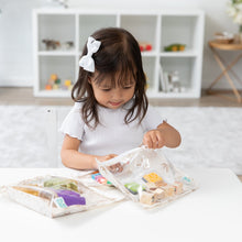 Load image into Gallery viewer, A young girl examines toys in a Bumkins Clear Travel Bag 3 Pack: Palm Daze at a white table in a bright room, wearing a white bow.
