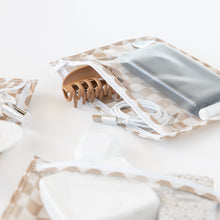 Load image into Gallery viewer, Close-up of Roamio by Bumkins Clear Travel Bag 3 Pack: Sand Check holding a hair clip, USB cable, and personal items on a white background.
