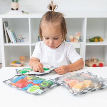 A child with a small ponytail sits at a white table with Bumkins Clear Travel Bag 3 Pack: Charcoal Check filled with toy blocks.