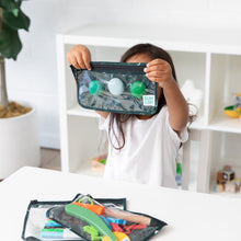 Load image into Gallery viewer, A child inspects items in a Bumkins Clear Travel Bag 3 Pack: Starry Cactus at a white table, while a plant adds a natural touch.
