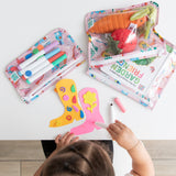 A child draws on colorful craft paper at a white table with Bumkins: Clear Travel Bag 3 Pack: Boots nearby filled with toys and markers.